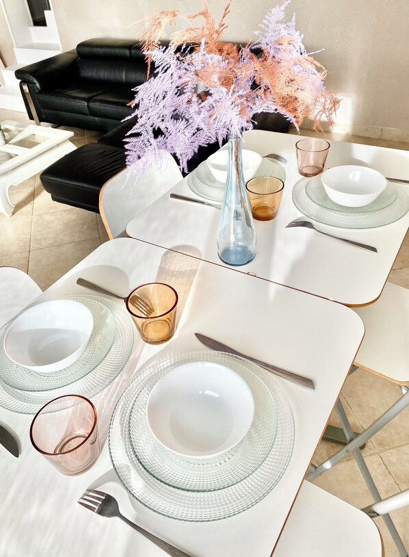 Une table à manger dressée avec de l’argenterie blanche, de la verrerie transparente et un vase contenant des fleurs séchées.