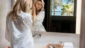 A person in a white robe standing in front of a bathroom mirror with a view of palm trees and a body of water outside. - Twinpalms MontAzure Phuket (Phuket)