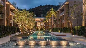 A modern hotel with a central pool, surrounded by lush greenery and a mountainous backdrop.