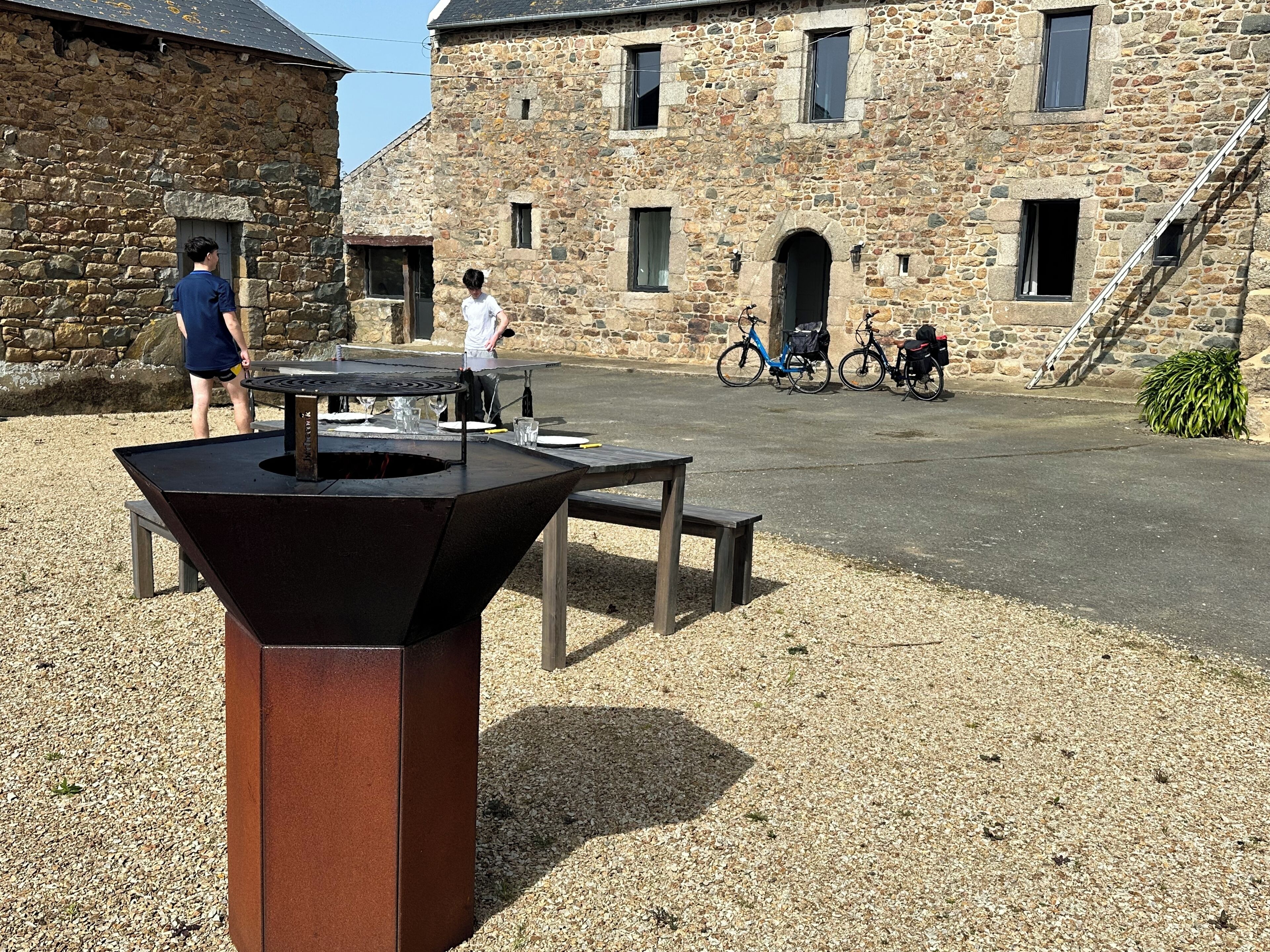 An outdoor area with a barbecue grill, picnic table, and bicycles parked near a stone building.