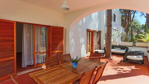 A wooden dining table with chairs, a potted plant, and a sofa in an outdoor patio area with wooden shutters and a view of trees.