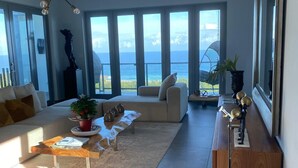 Een ruime woonkamer met een groot raam met uitzicht op zee, een moderne kroonluchter en een houten salontafel.