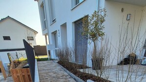 Ein gepflasterter Weg mit einem Entwässerungsrost, flankiert von Pflanzkästen mit Sträuchern und einem Holzzaun.