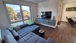 Ein modernes Wohnzimmer mit einem großen, grauen Ecksofa, einem Flachbildfernseher, der an einem hellblauen Schrank befestigt ist, und einem Essbereich mit einem Holztisch und Stühlen.