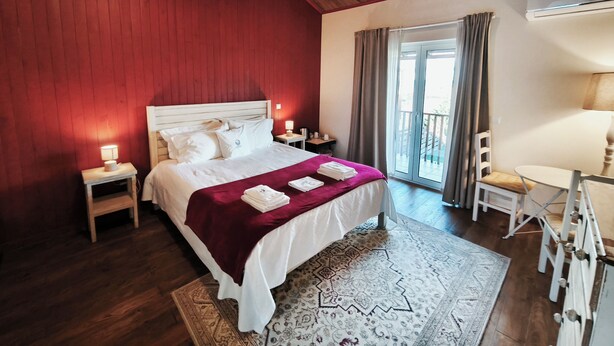 Ein Hotelzimmer mit Holzboden, einem Bett mit weißer Bettwäsche und einer roten Decke, einem kleinen Nachttisch mit Lampe, einem Sessel, einem Schreibtisch mit Lampe und Blick auf einen Balkon durch eine Glastür.