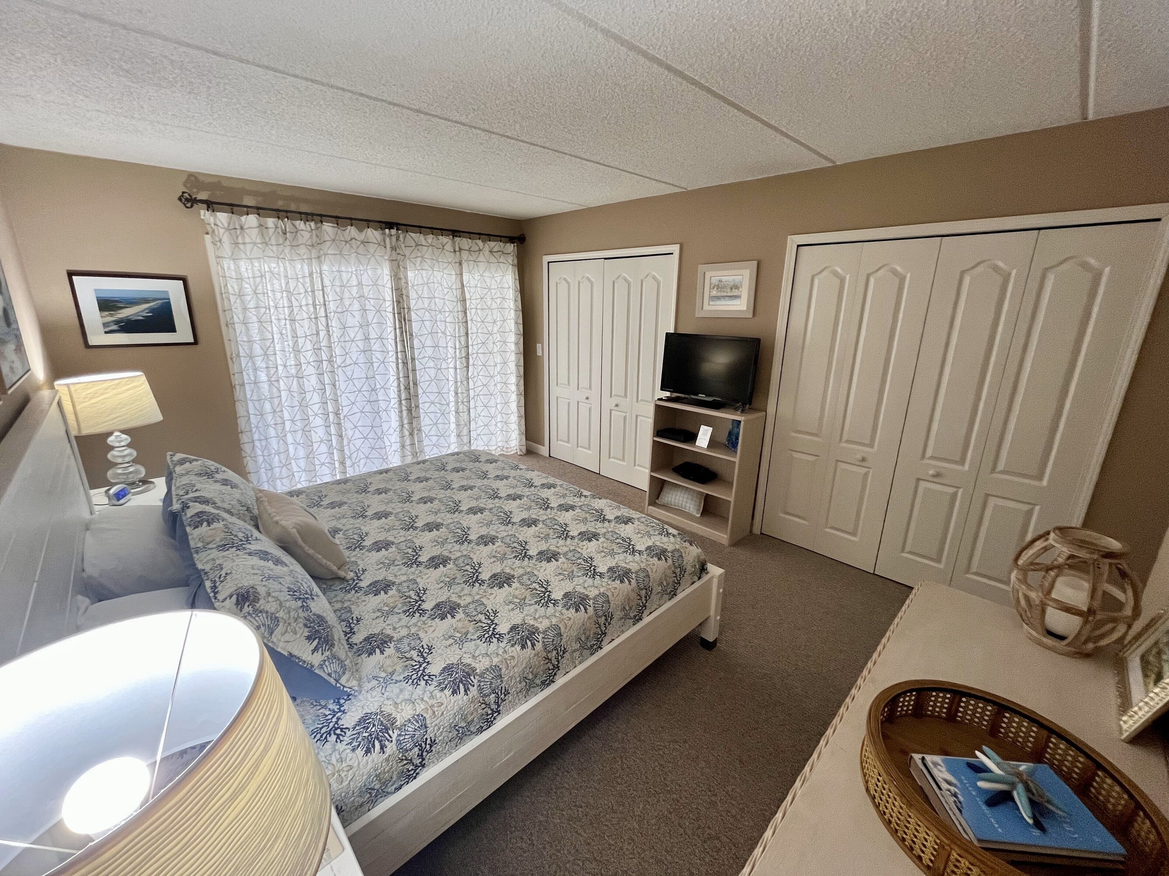 A bedroom with a bed, a TV on a stand, a lamp, a window with curtains, and a wardrobe.