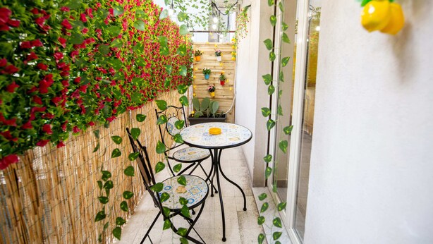 A narrow outdoor corridor with a small table and chairs, a bamboo fence with red flowers, and a wooden wall with potted plants. - Terra d'Agrumi Rooms (Catania)