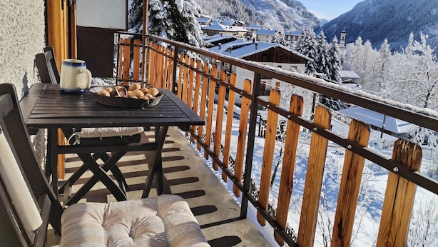 Una terraza de madera con mesa y sillas, un comedero para pájaros y vistas de montañas nevadas y edificios.