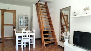 Una sala de estar con una escalera de madera, una mesa de comedor con sillas, un televisor sobre un mueble blanco y un espejo grande.