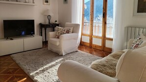 Una sala de estar con un televisor, un sofá blanco, una mesita con una lámpara y vistas a montañas nevadas a través de un ventanal.