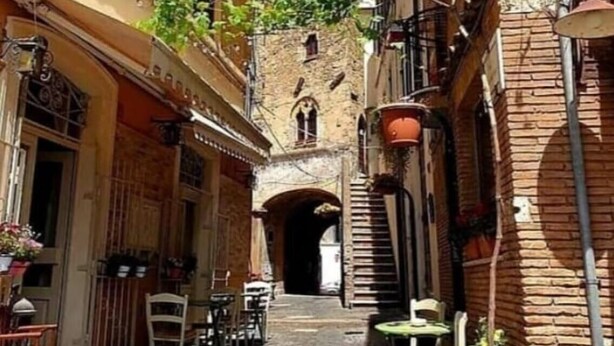 A narrow street with buildings on both sides, a stone archway, and outdoor seating with tables and chairs. - Alice's House a stone's throw from the sea and the old town of Neptune (Nettuno)