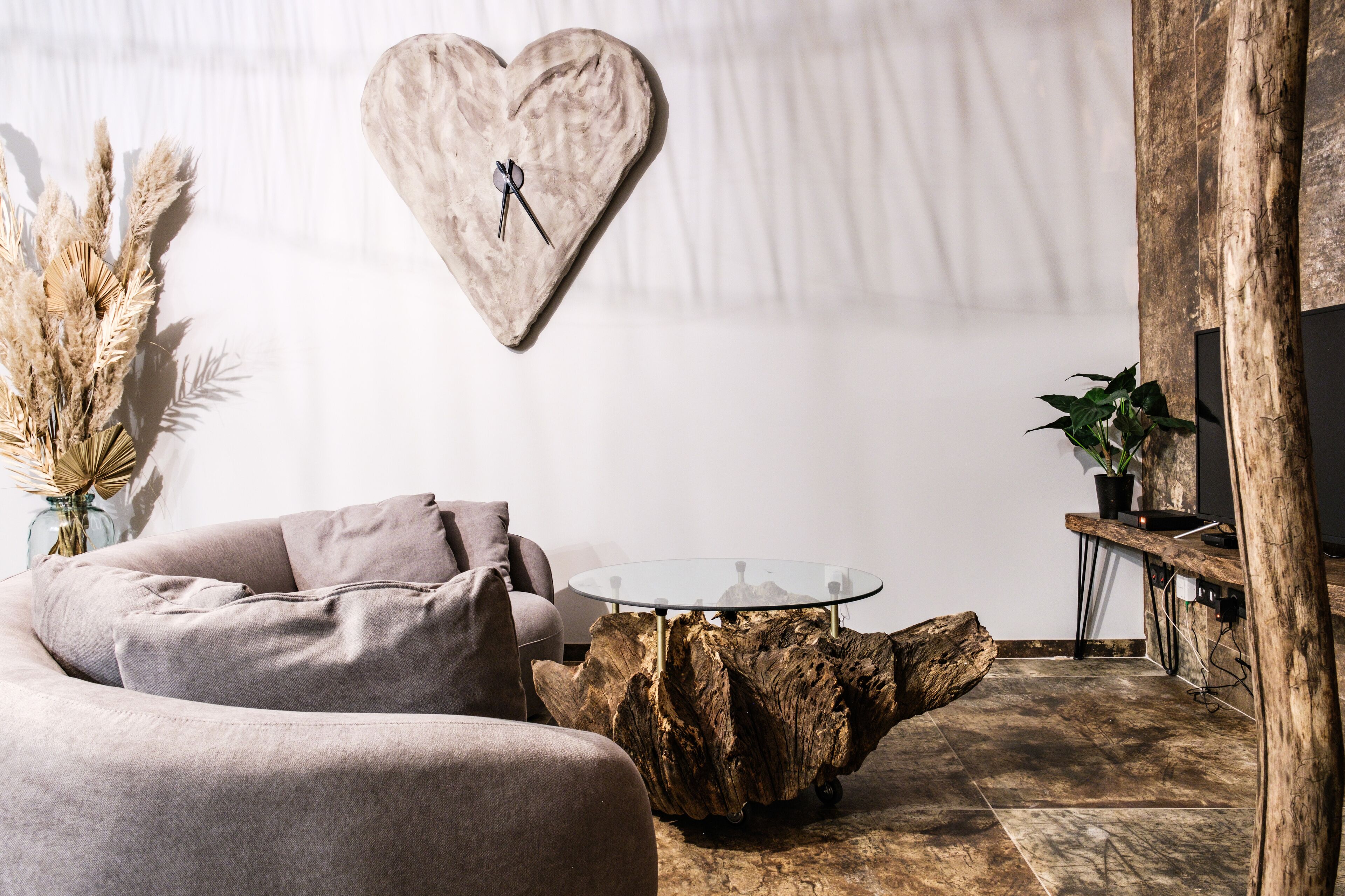 Un salon moderne avec une table basse ronde en verre, un élément décoratif mural en forme de cœur, une table d’appoint en bois rustique et un canapé.
