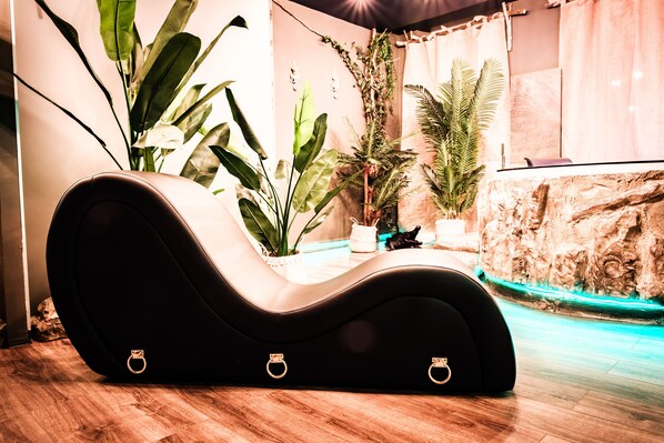 A curved black lounge chair with gold rings, surrounded by large green plants and a rock wall.