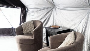 Quarto com tenda, dois poltronas, uma mesinha pequena, soalho de madeira e uma janela.