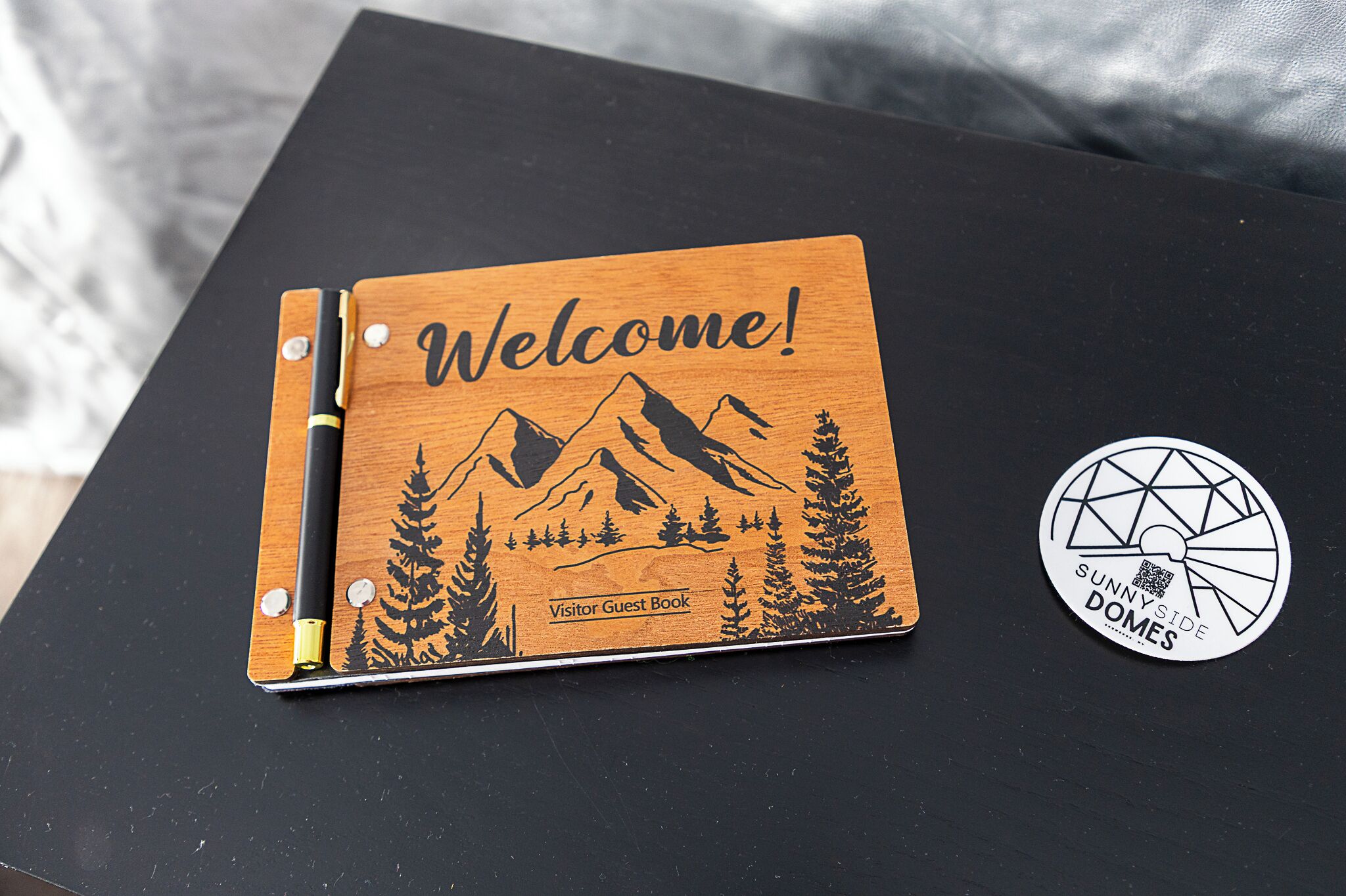 A wooden welcome sign with a mountain and tree design, labeled "Visitor Guest Book," and a circular sticker with "Sunny Side Domes" text.