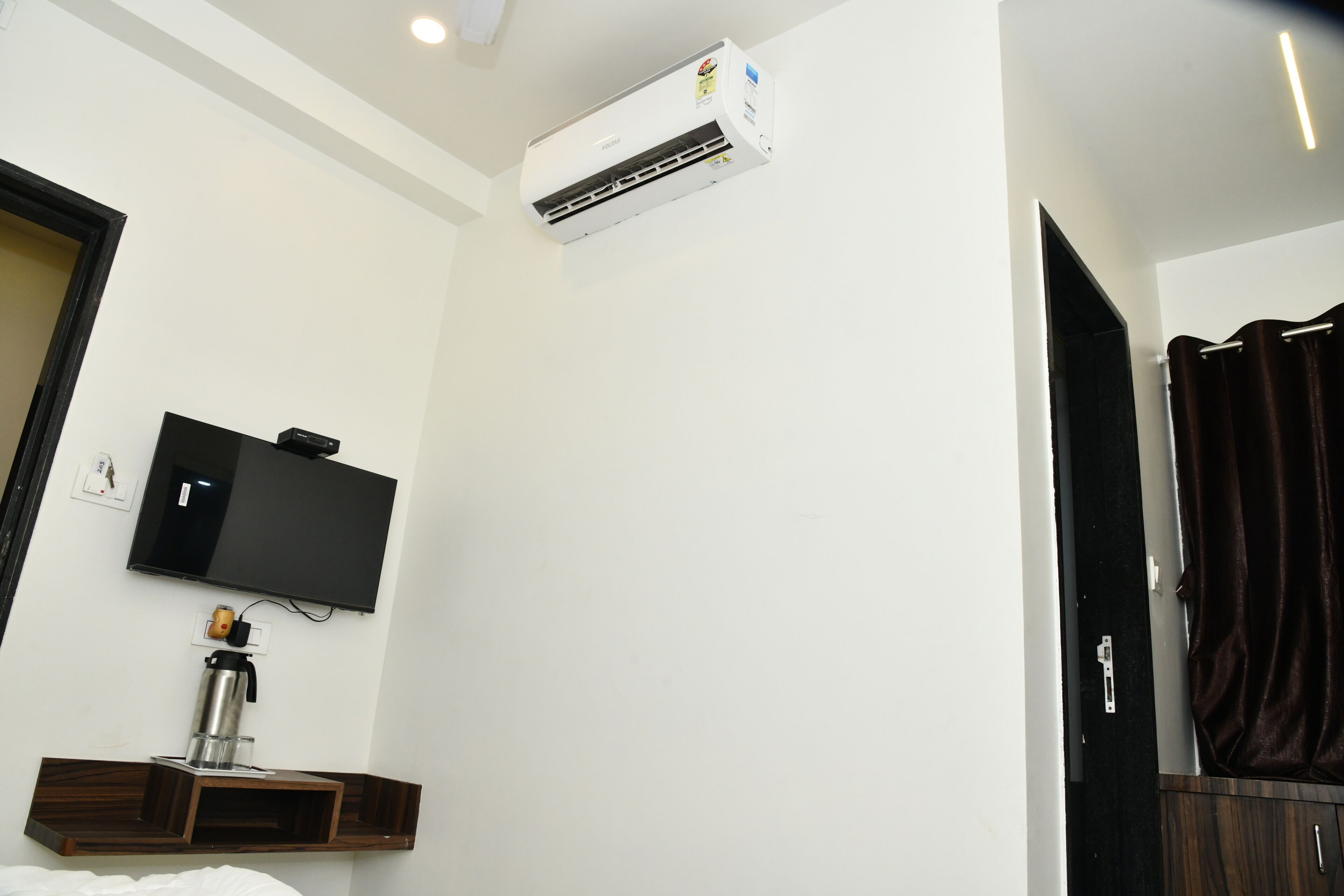 Habitación de hotel con aire acondicionado montado, televisor de pantalla plana, mesita de noche con cafetera y cortina oscura.