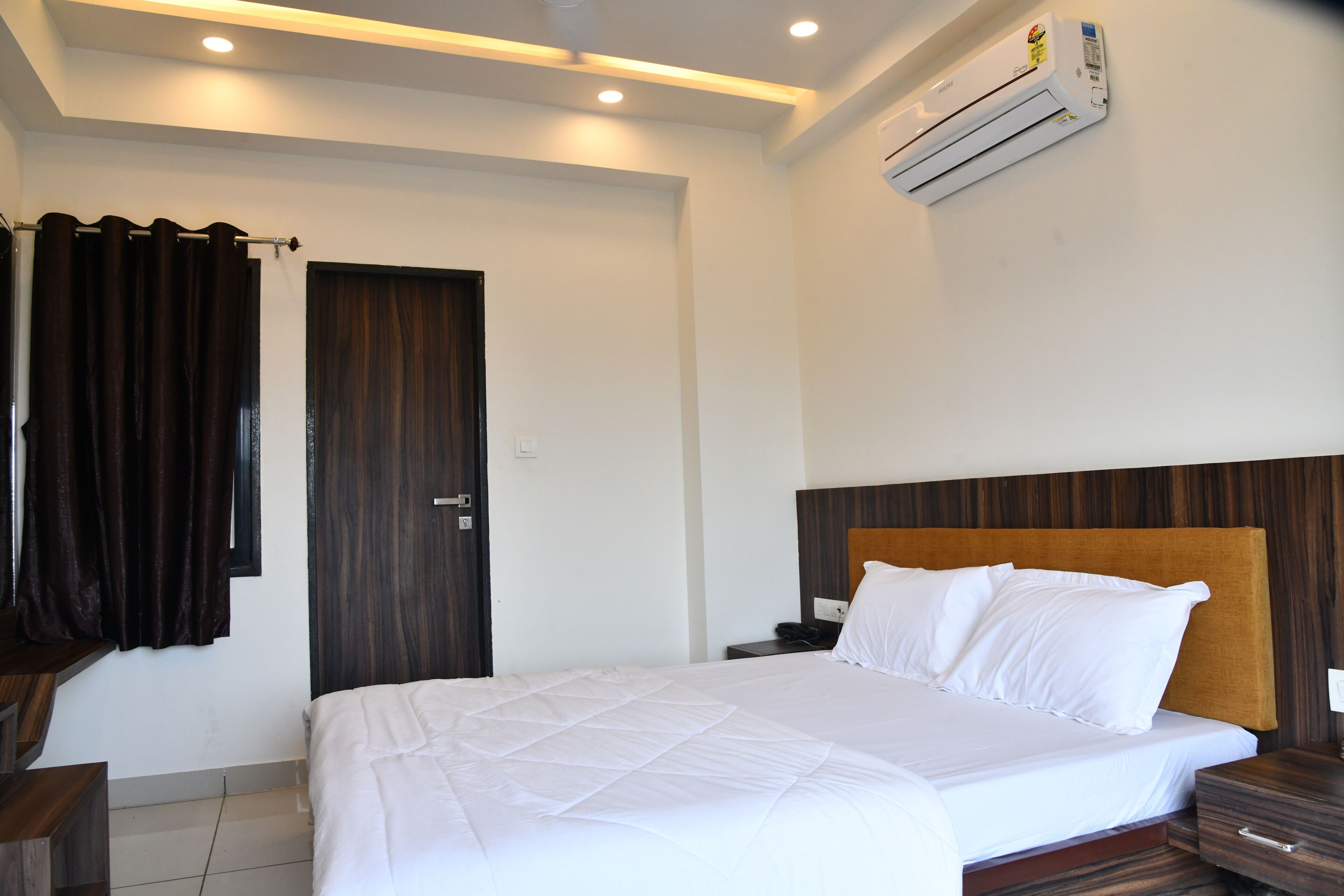 Habitación de hotel con una cama grande, cabecera de madera, mesita de noche y un equipo de aire acondicionado, junto a una puerta con cortinas.