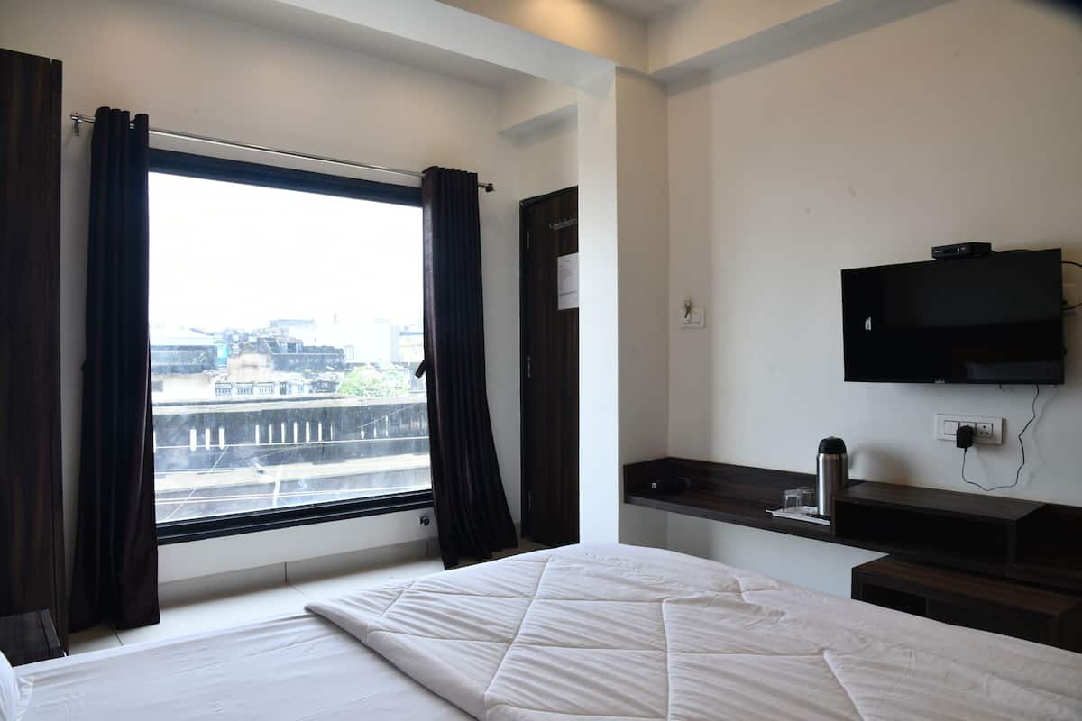 Quarto de hotel com uma janela grande, uma cama, uma televisão na parede e uma pequena mesa com uma chaleira e copos.