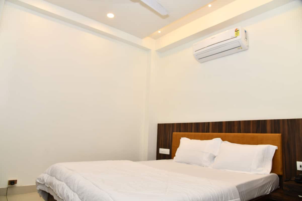 Quarto de hotel com uma cama de solteiro, cabeceira de madeira, roupa de cama branca e um aparelho de ar condicionado fixado na parede.