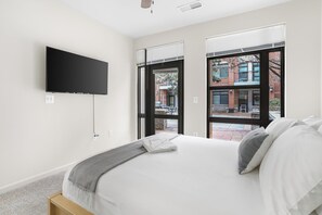 A bedroom with a bed, a flat-screen TV mounted on the wall, and large windows offering a view of the outside. - Crystal City Apartments near Crystal Drive - We Host (Arlington)