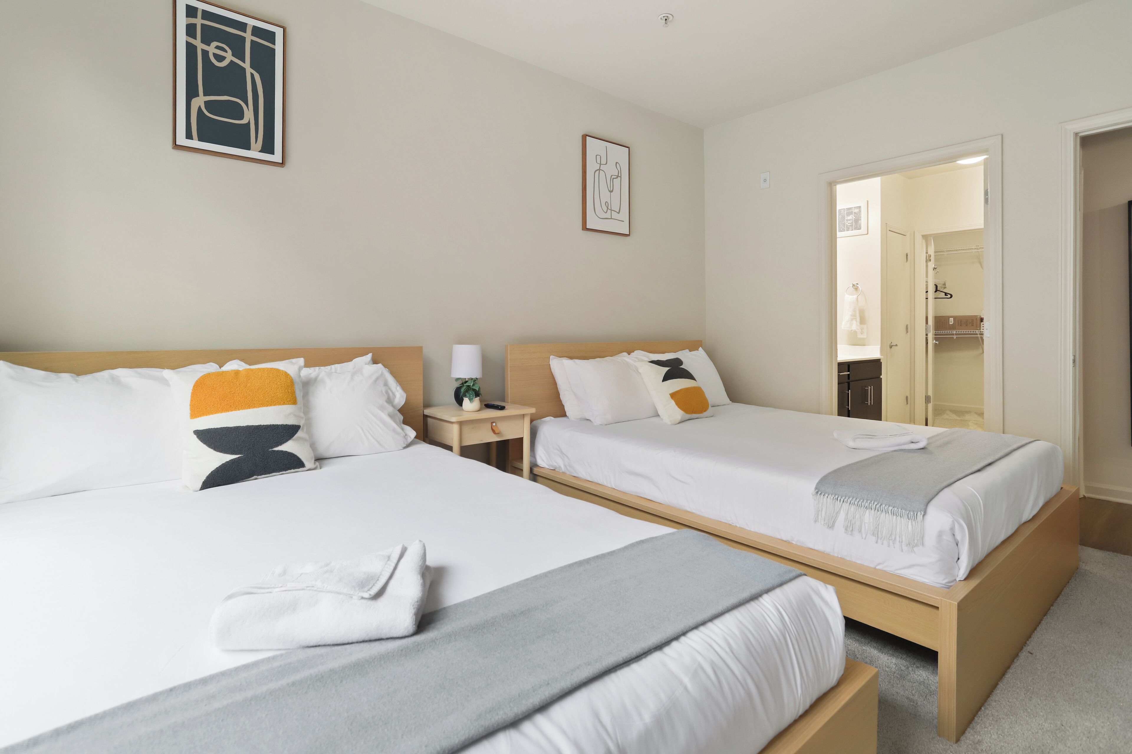 A hotel room with two beds, a bedside table with a lamp, a framed picture on the wall, and a closet with hanging clothes.