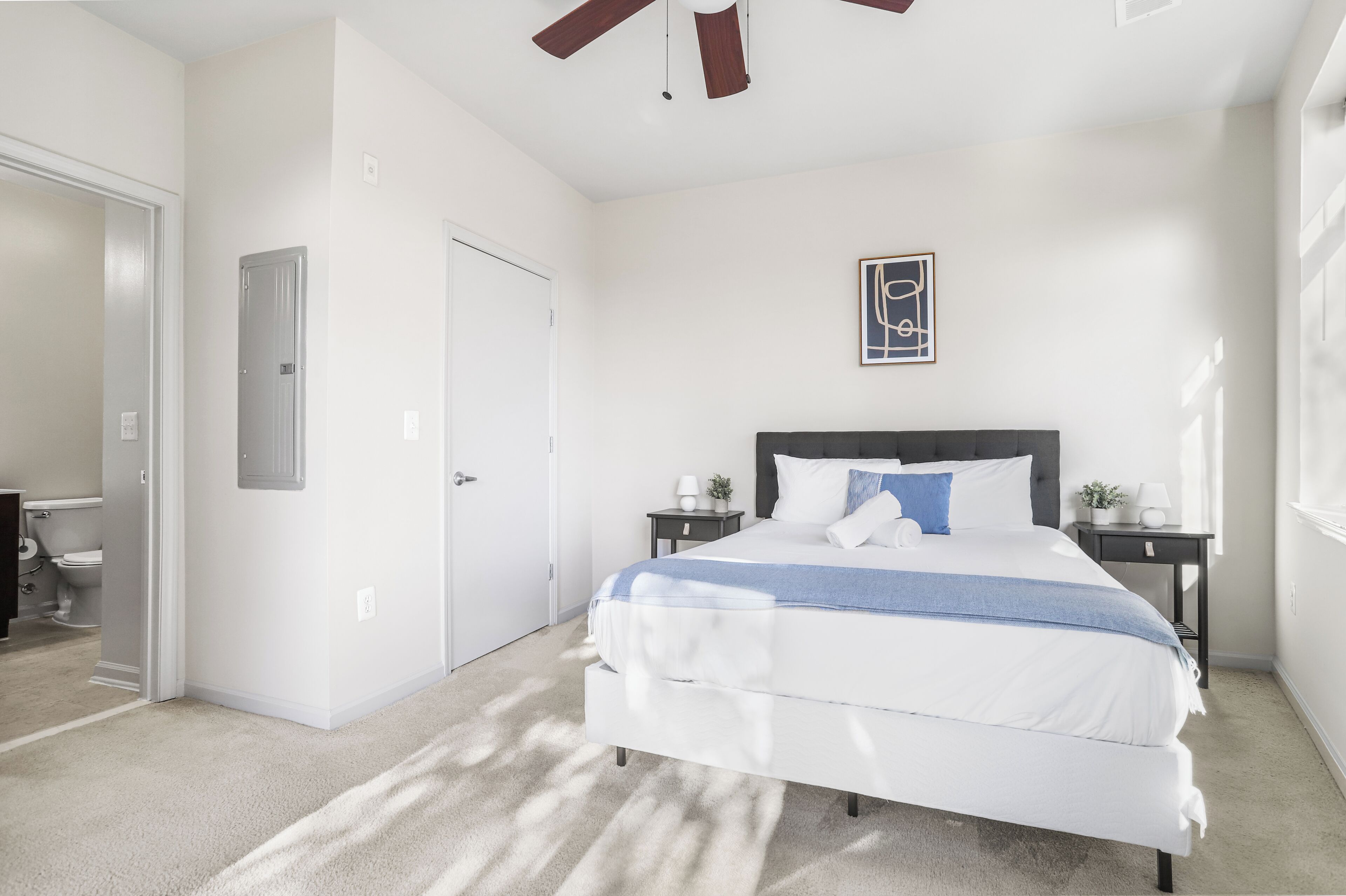 A bedroom with a large bed, a ceiling fan, a door, and a small bedside table with a lamp.