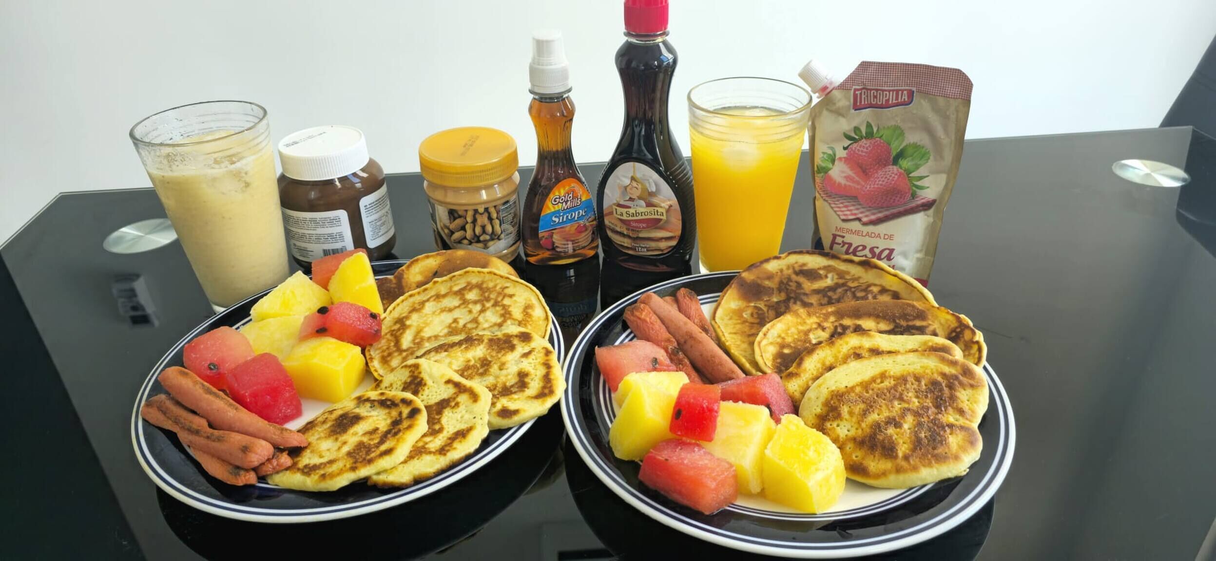 Two plates of pancakes with fruit and syrups, a bottle of honey, a glass of orange juice, and a carton of milk on a table.
