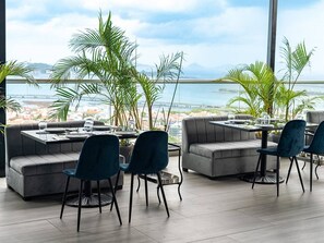 A modern dining area with grey sofas and blue chairs, large windows offering a view of the sea and cityscape. - Room-sofa Bed Stay Rooftop Pool & Gym, A (Panama City)