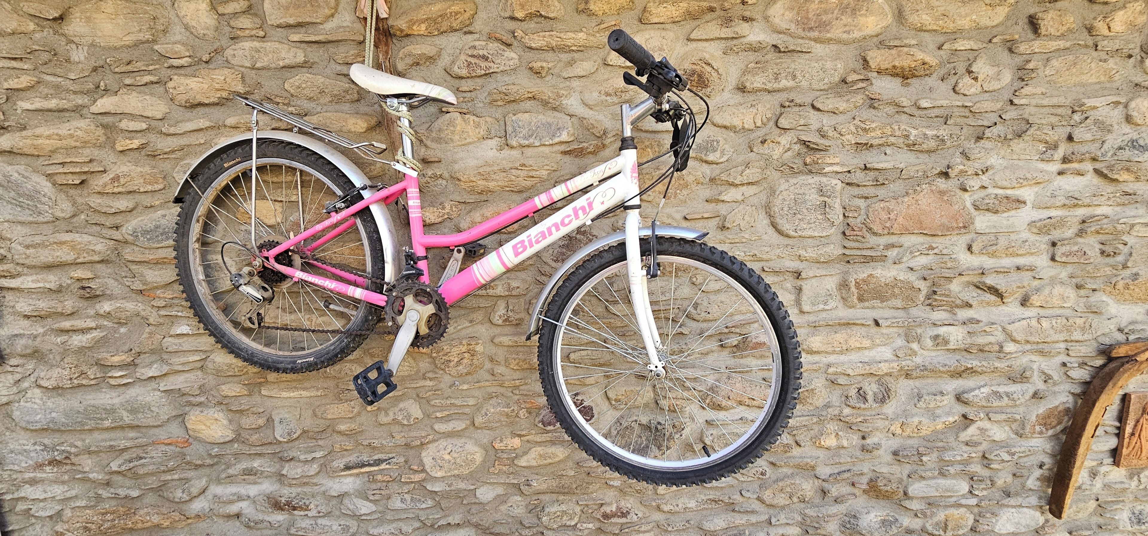 Ένα ροζ ποδήλατο με λευκή σέλα και τιμόνι, ακουμπισμένο σε έναν πέτρινο τοίχο.
