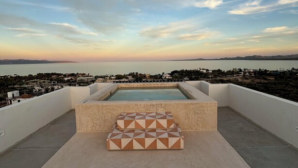 A rooftop terrace with a swimming pool, geometric-patterned cushions, and a view of the city and mountains. - Modern 2 bed/2 bath, 1st floor condo. 
Casa Ventosos (Calypso 102). (La Ventana)