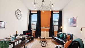 A modern living room with a dining area, featuring a large window with curtains, a clock, a Netflix display, and a cozy sofa with cushions. - The Jewel Box 3BR Apt, JQ Birmingham with Parking (birmingham)