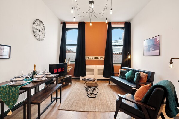 Een moderne woonkamer met een eethoek, met een groot raam met gordijnen, een klok, een Netflix-scherm en een gezellige bank met kussens.