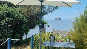An outdoor table and chairs with a white umbrella, surrounded by greenery and overlooking a body of water.