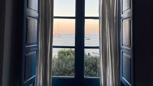 A room with a large window featuring white curtains, a view of the sea, and a sailboat in the distance.