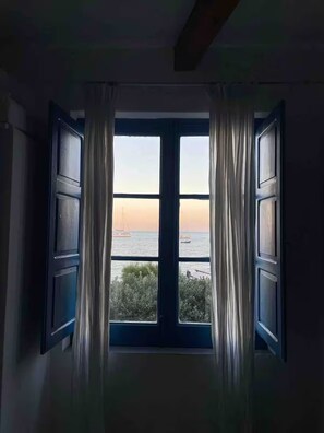 A room with a large window featuring white curtains, a view of the sea, and a sailboat in the distance. - Mini apartment with charming window overlooking the sea quiet and relaxing. (Lipari-Stromboli)