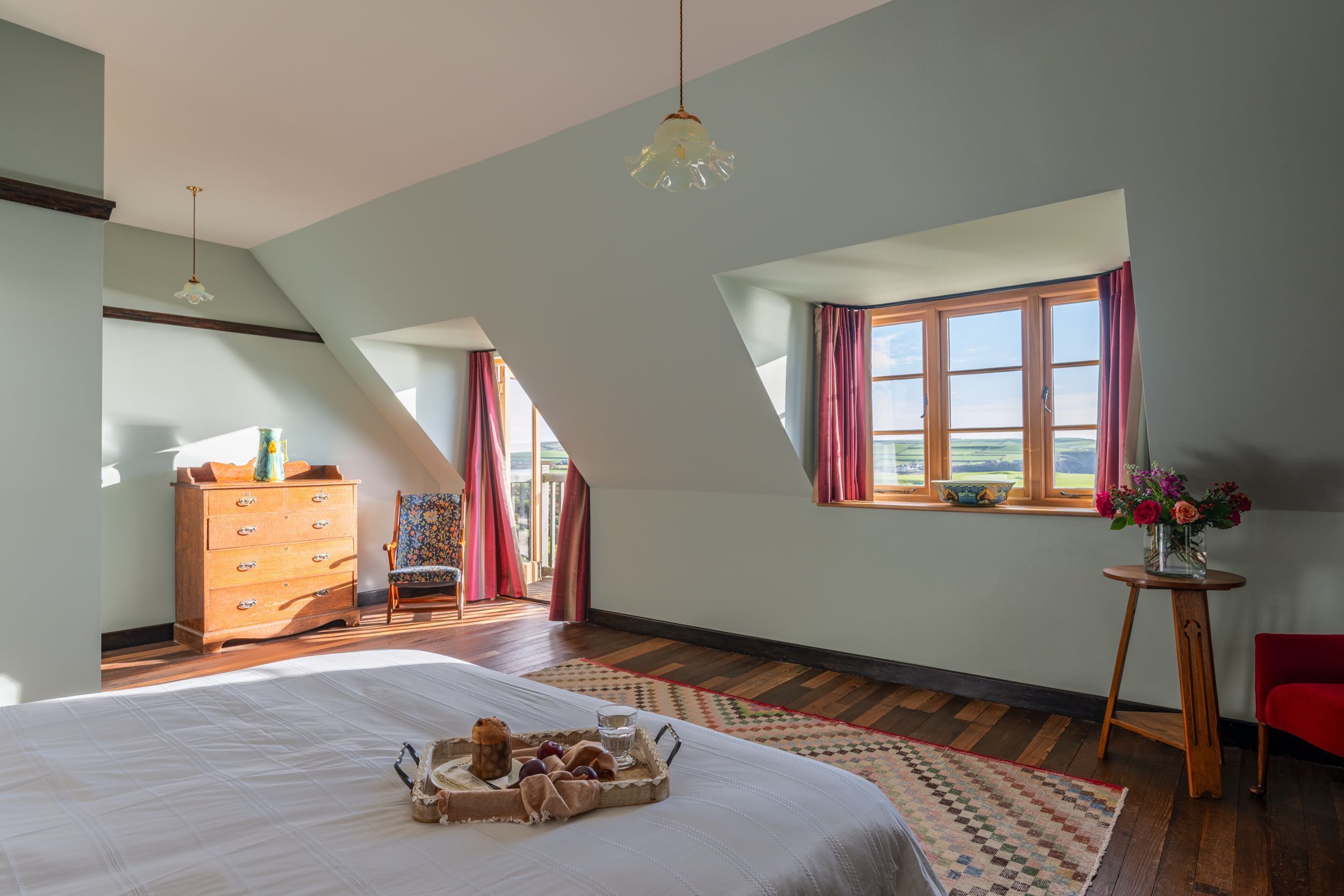 Ein Schlafzimmer mit einem großen Bett, einem Schminktisch, einem Sessel, einem kleinen Tisch mit Blumen und einem Fenster mit Vorhängen.