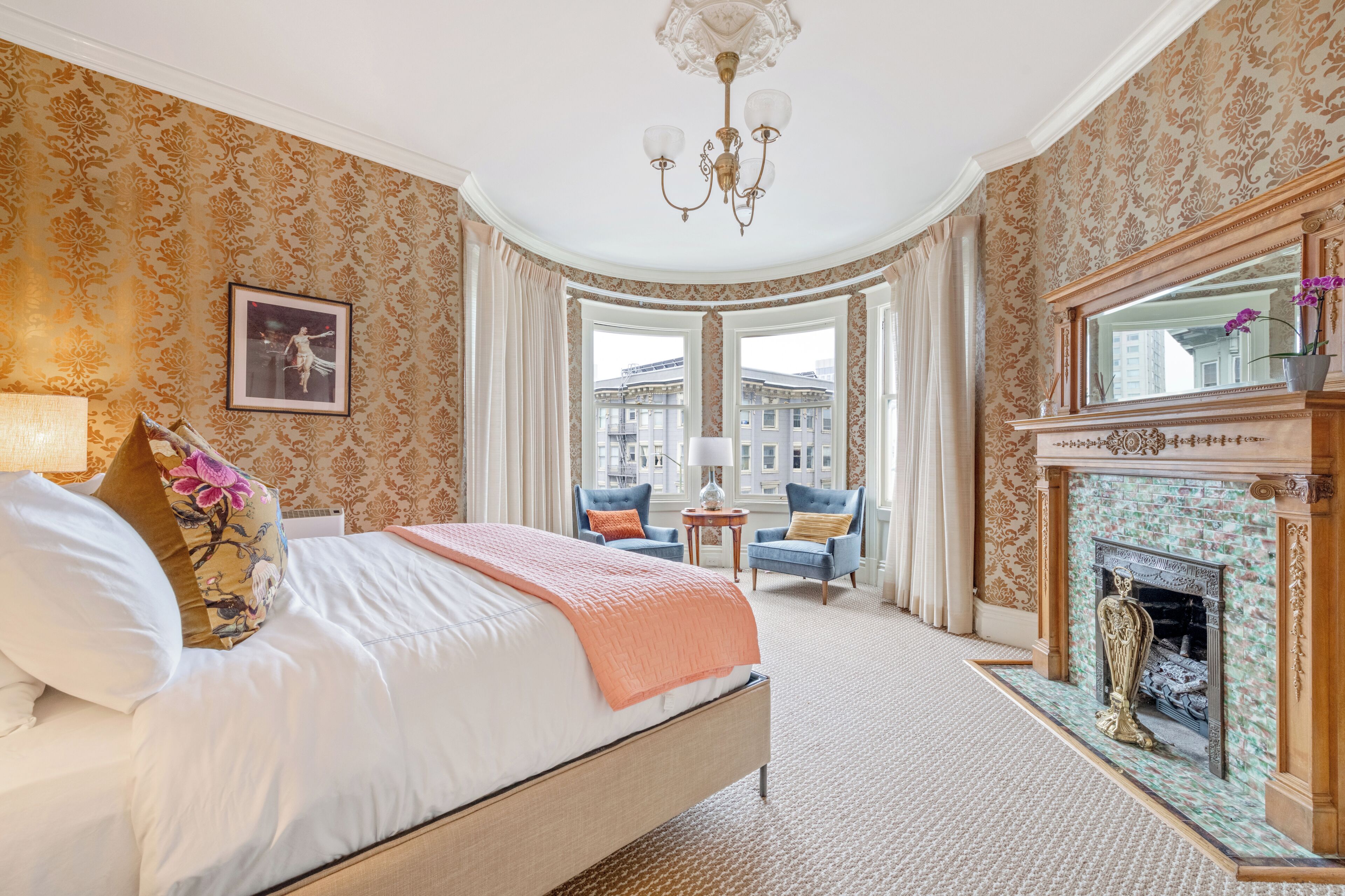 A hotel room with a large bed, a fireplace, a view of buildings, and a chandelier.
