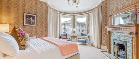 A hotel room with a large bed, a fireplace, a view of buildings, and a chandelier.