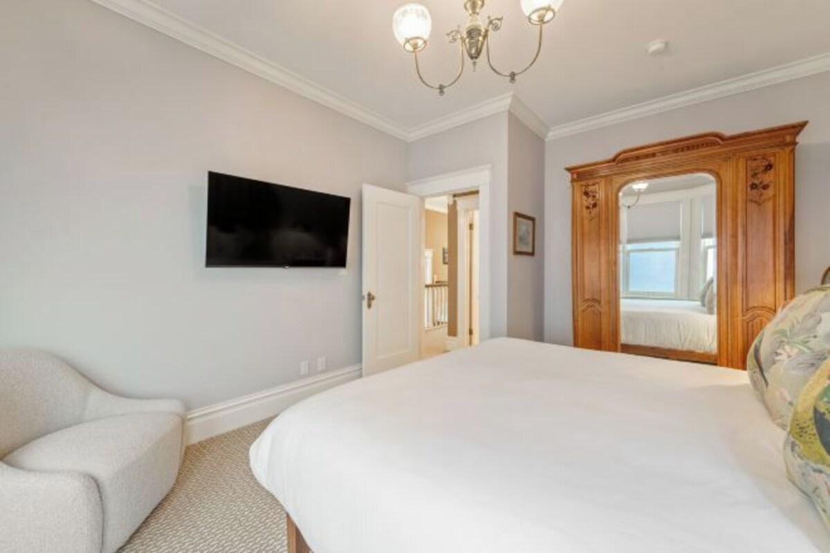 A bedroom with a large bed, a television mounted on the wall, a wooden wardrobe, and a door leading to another room.