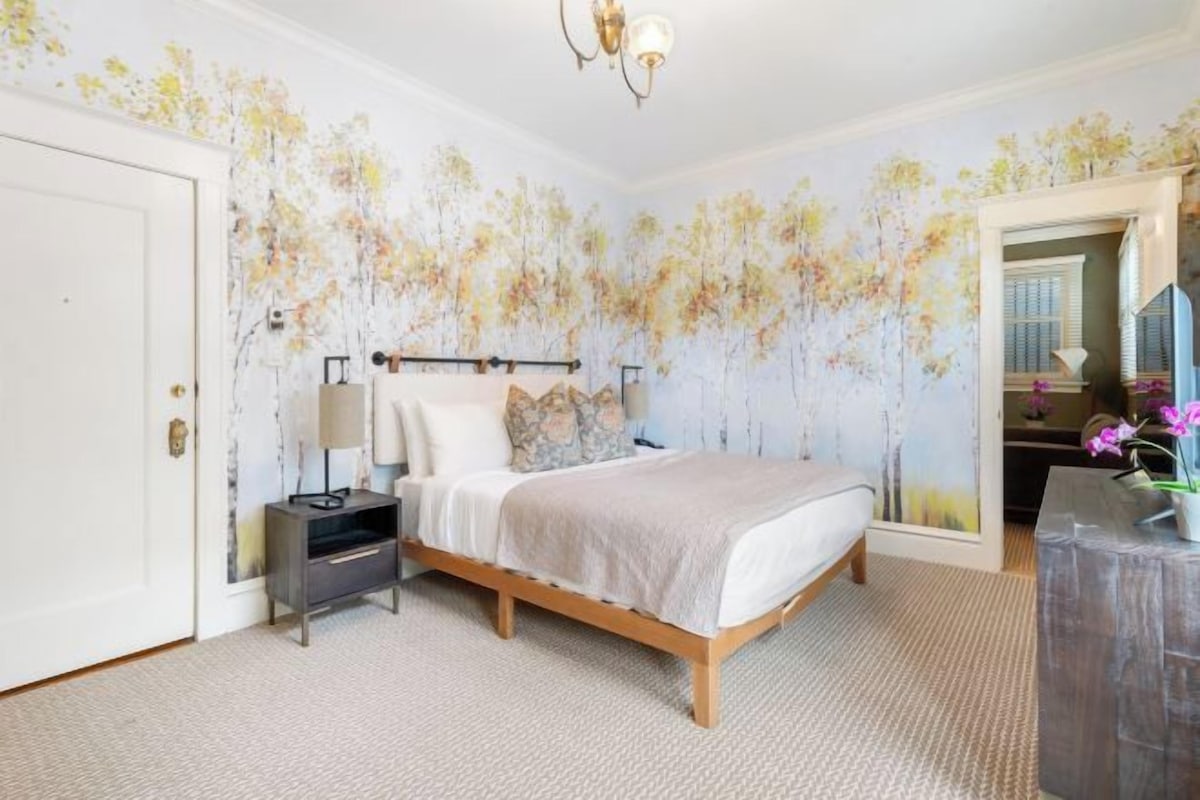 Une chambre à coucher comprenant un lit, une table de chevet, une lampe et un mur orné d’une fresque florale.