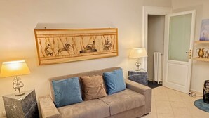 A living room with a beige sofa, two blue cushions, a marble side table, a wall-mounted lamp, and a large framed artwork.