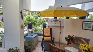 En patio med et træbord, stole, en gul parasol, plantekasser og en fiskedekoration på væggen.