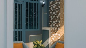 Une pièce avec une porte bleue, deux fauteuils en osier dotés de coussins orange, une petite table sur laquelle est posée une plante, et un mur orné d’un motif.