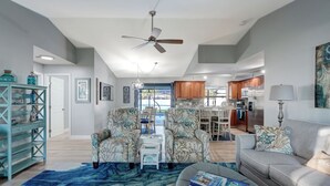 Ein geräumiges Wohnzimmer mit Deckenventilator, einem grauen Sofa und gemusterten Sesseln. Im Hintergrund ist eine Essecke mit Holzausbau und Blick ins Freie zu sehen.