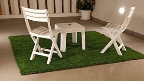 Eine Terrassenfläche mit zwei weißen Stühlen und einem kleinen Tisch auf künstlichem Rasen, umgeben von einem Betonboden und einem weißen Geländer.