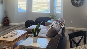 A spacious living room with a ceiling fan, a large clock, and a view through the windows. - Lake Powell, Grand Circle, 3-bedroom house with AC, WiFi in welcoming Big Water (Big Water)
