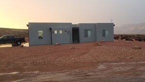 A modern building with a flat roof, a door, and windows, situated in a desert with sparse vegetation and a mountain in the background. - Lake Powell, Grand Circle, 3-bedroom house with AC, WiFi in welcoming Big Water (Big Water)
