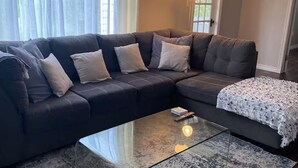 Un salon avec un canapé d’angle en cuir foncé, une table basse en verre et un tapis à motifs.
