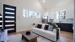 A modern living room with a white sofa, a wooden coffee table, and a black bowl. There is a kitchen area with dark cabinets and a refrigerator. - Cozy/Modern King Suite in Houston Texas (Houston)