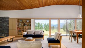 A modern living room with a stone wall, wooden ceiling, and large windows offering a view of trees and mountains. - Contemporary Berkshire Home with Views of Taconic Golf Course (Williamstown)